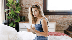Woman holding up menstrual cup and reading info card about it while sitting in a bedroom with plants, sustainable menstrual products, menstrual cup, menstrual cups, period cup, june menstrual cup